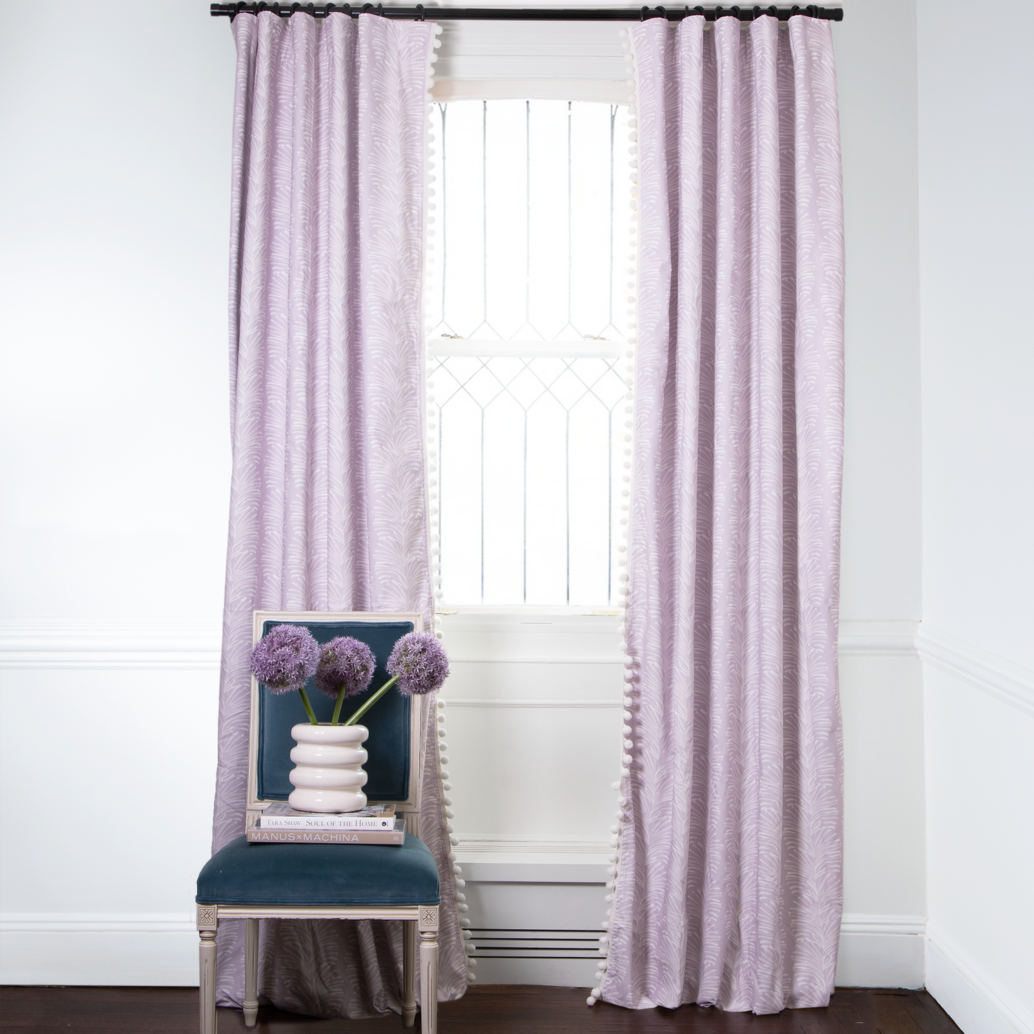 Lavender Botanical Stripe Printed curtains with White Pom Poms on metal rod in front of an illuminated window with Navy Velvet chair with Purple Flowers in white vase on top of two stacked books