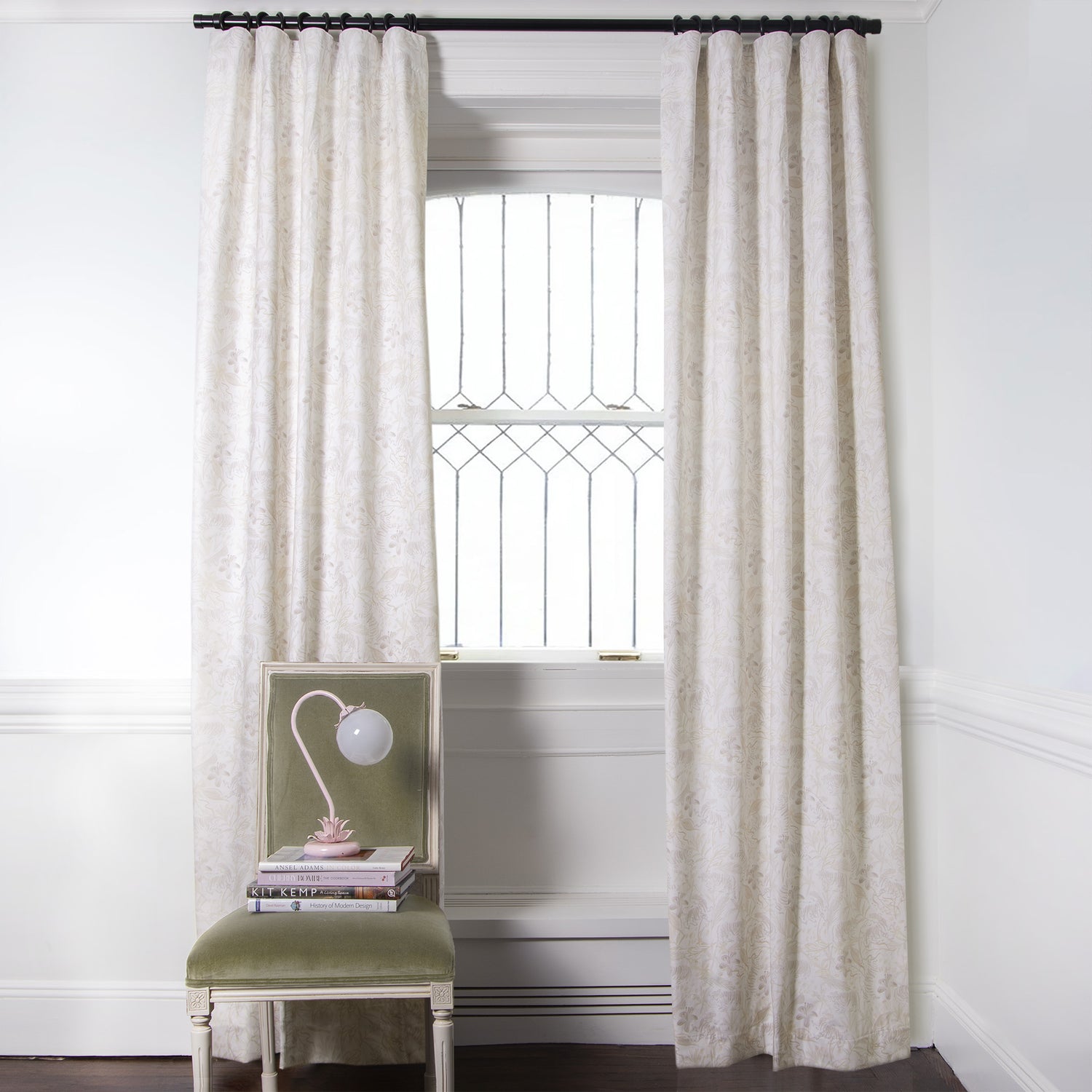 Beige Chinoiserie Tiger Printed Curtains on metal rod in front of an illuminated window with Green Velvet chair with floral pink lamp and stacked books on top