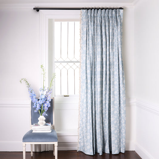 Cornflower Blue Floral Printed Curtains on metal rod in front of an illuminated window with Baby Blue Velvet chair with blue flowers in white vase stacked on books