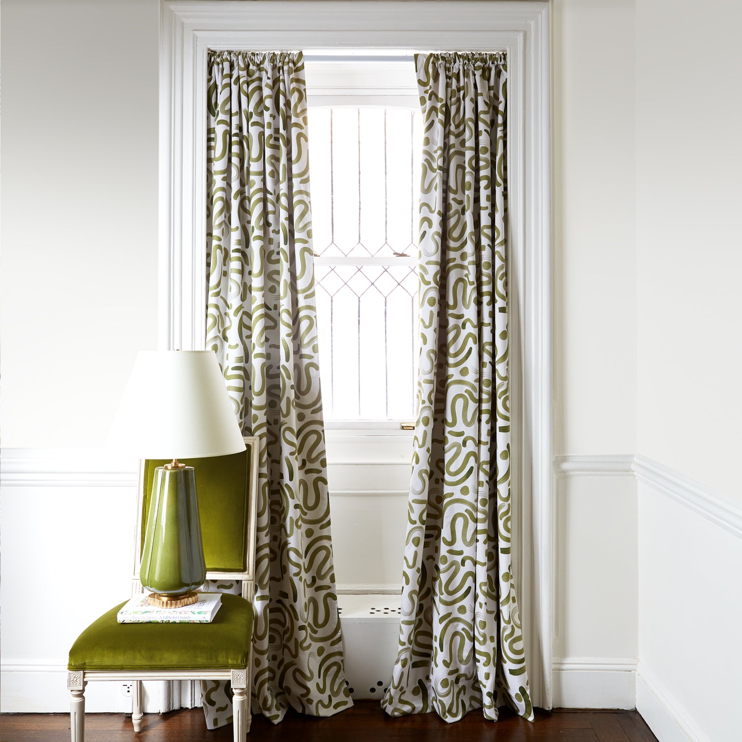 Moss Green Printed Curtains on white rod in front of an illuminated window with Green Velvet chair with white and green lamp on top of white book