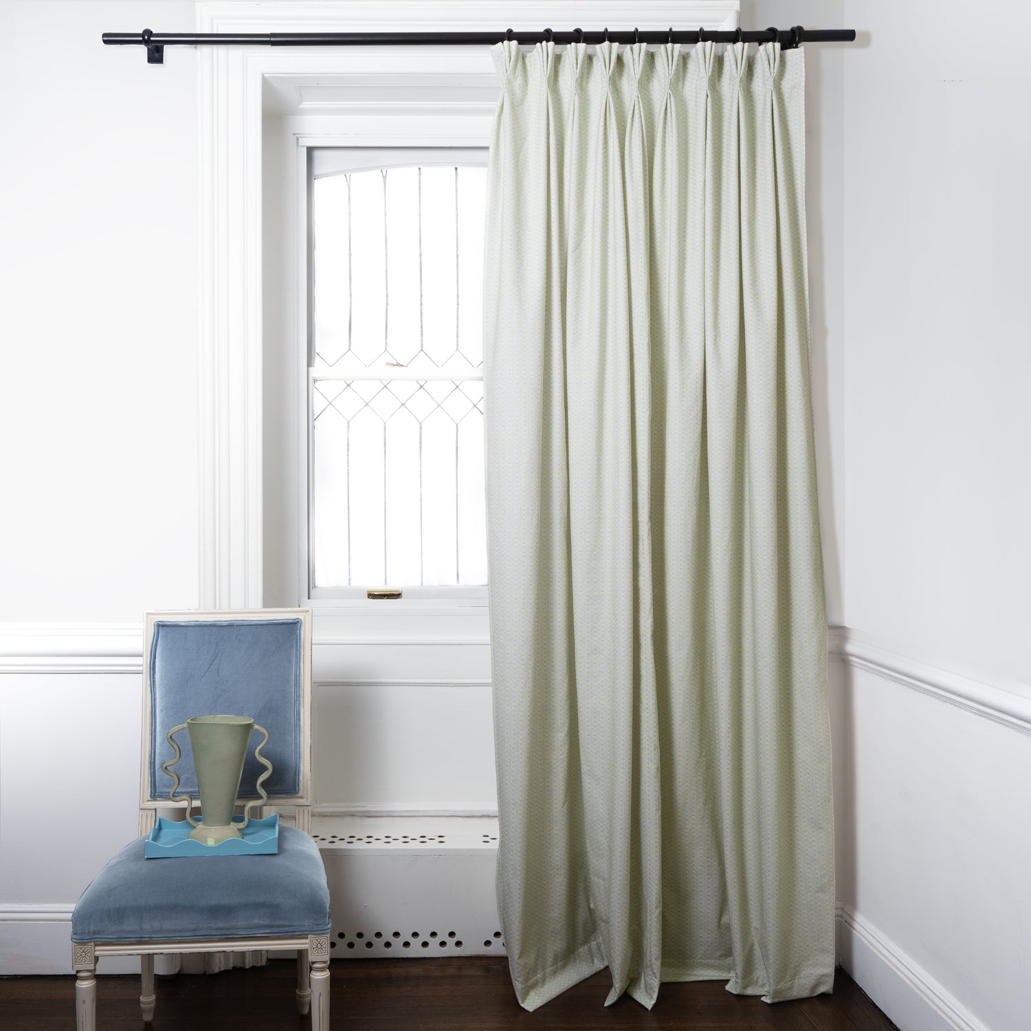 Moss Green Geometric Printed Curtains on metal rod in front of an illuminated window with Blue Velvet chair with green vase on top of blue plate