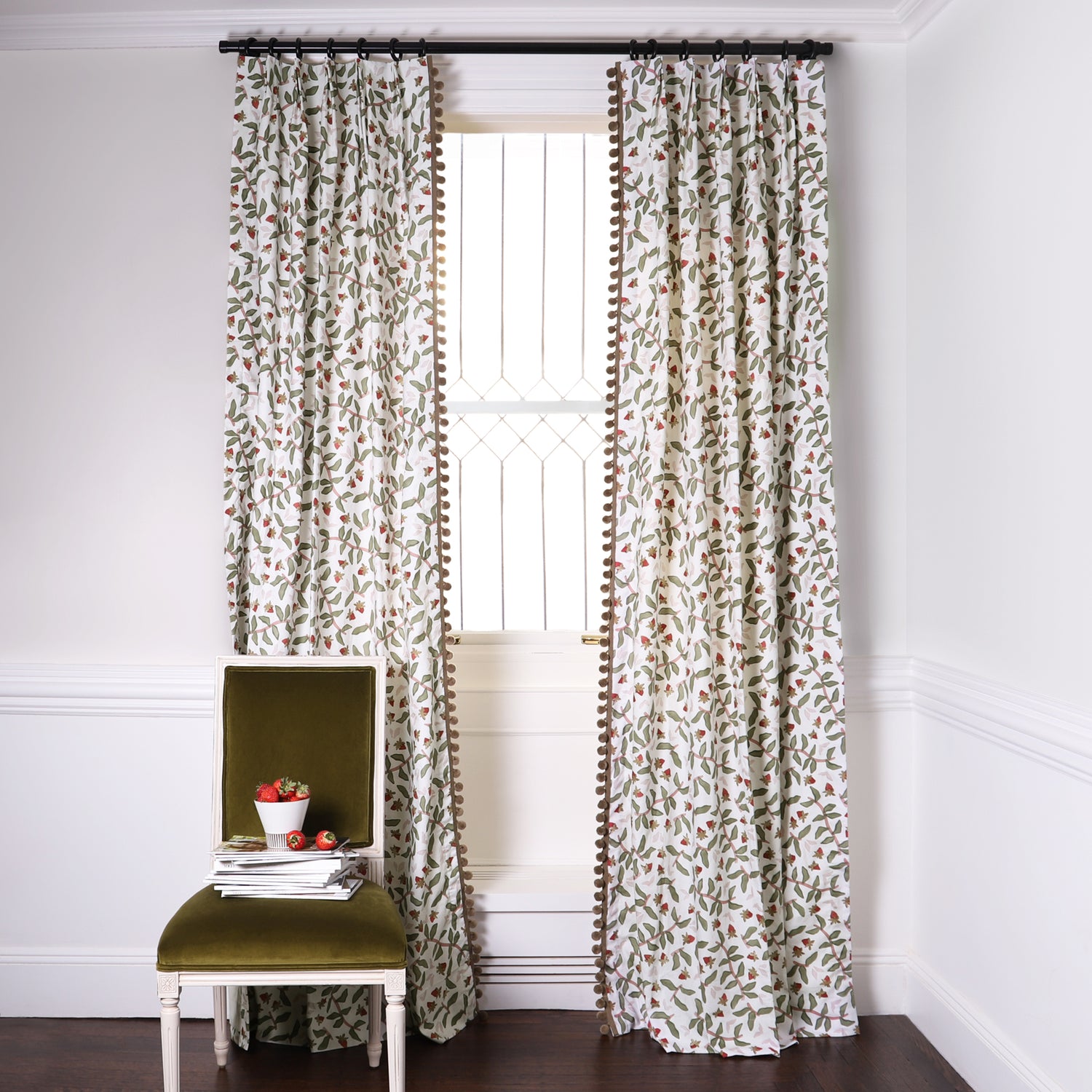 Strawberry & Botanical Printed Curtains with sage green pom pom on metal rod in front of an illuminated window with Moss Green Velvet chair with strawberries in white bowl on top of stacked magazines