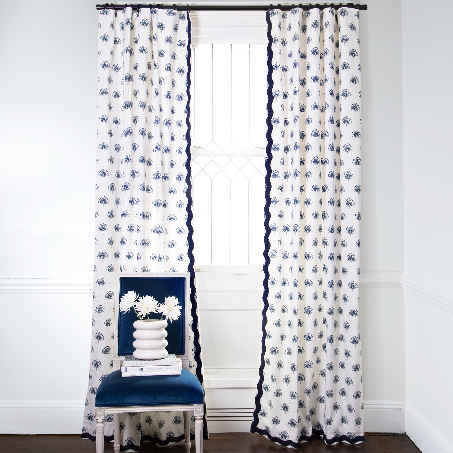 Floral Navy Printed curtains with Navy trim on metal rod in front of an illuminated window with Blue Velvet chair with white flowers in white vase on top of stacked books