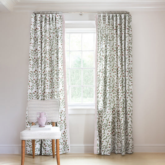 Strawberry & Botanical Printed Curtains with sage green pom pom on metal rod in front of an illuminated window with Moss Green Velvet chair with strawberries in white bowl on top of stacked magazines