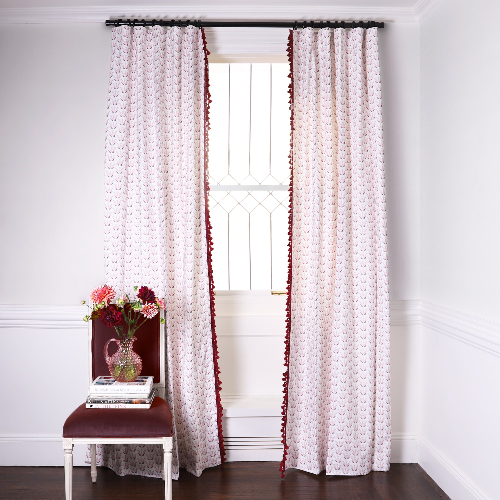 Pink & Burgundy Drop Repeat Floral Printed Curtains on black rod in front of an illuminated window with a burgundy Velvet chair with plants in clear pitcher vase on top of stacked books