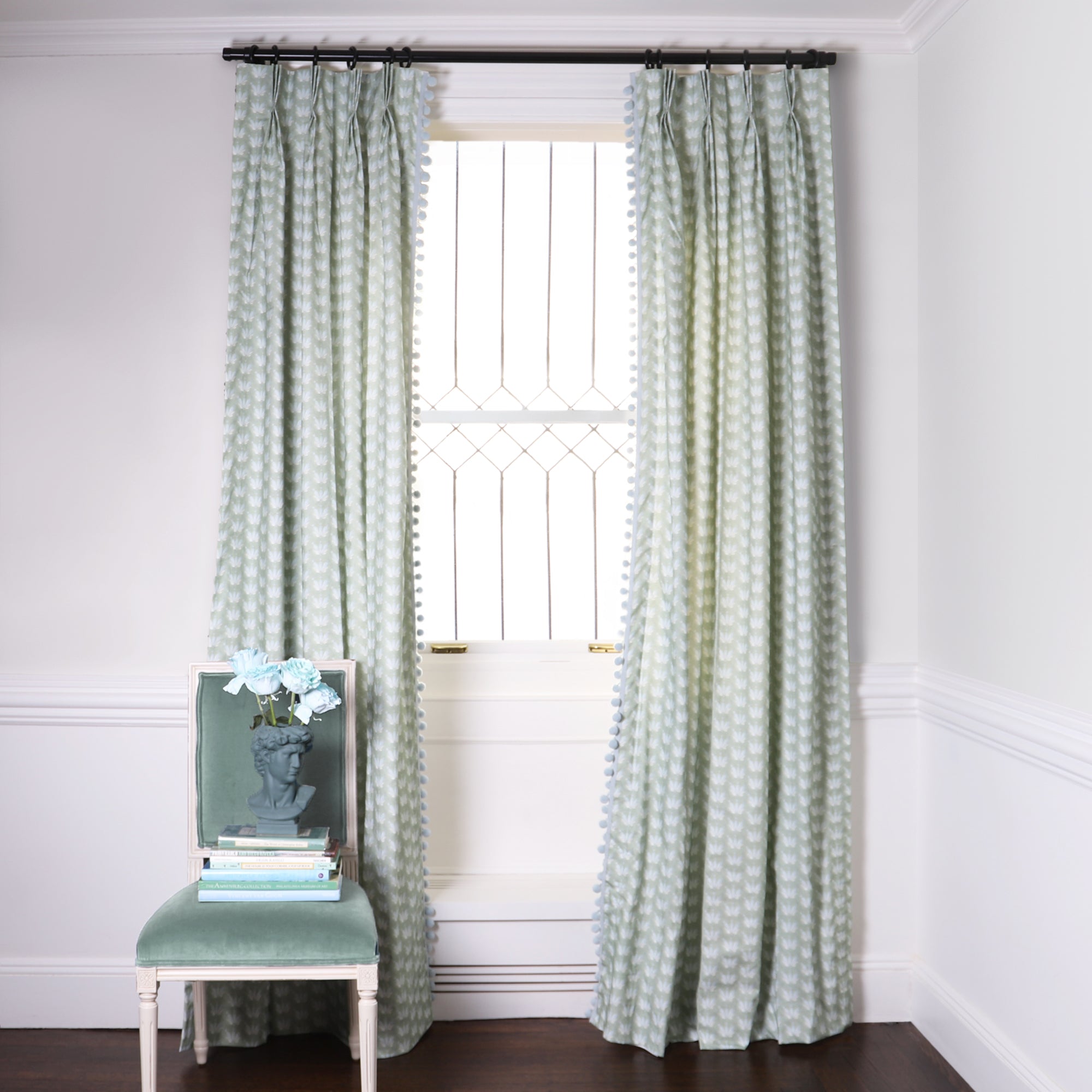 Blue & Green Floral Drop Repeat Printed Curtains on black rod in front of an illuminated window with a Blue Green Velvet chair with blue flowers in blue sculpted vase on top of stacked books