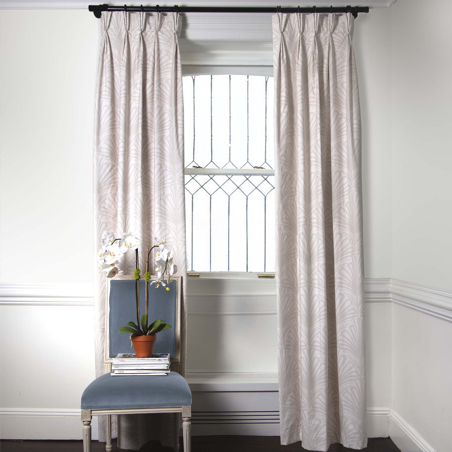 Beige Palm Printed Curtains on metal rod in front of an illuminated window with sky blue velvet chair and white flowers in clay pot on stacked books