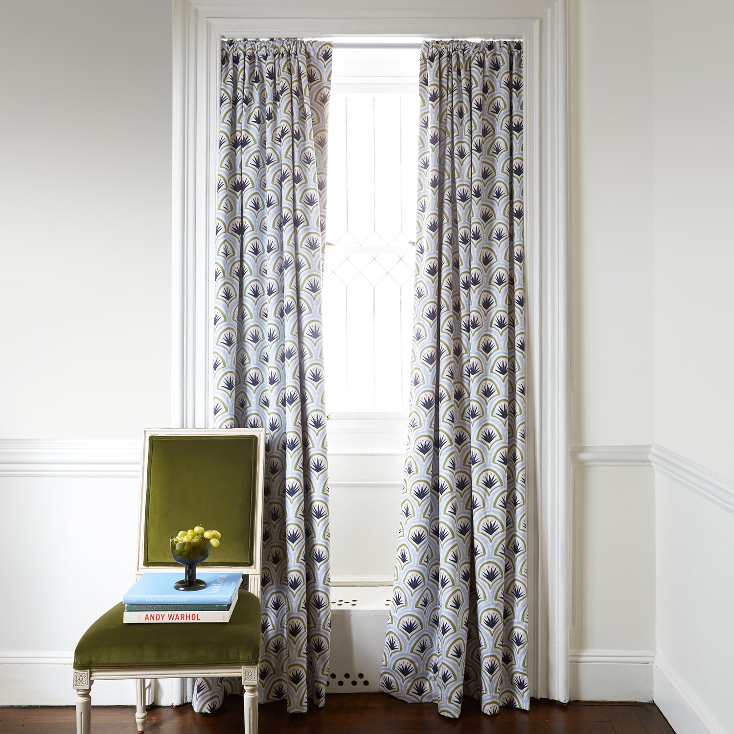 Art Deco Palm Pattern Printed Curtains on white rod in front of an illuminated window with Green Velvet chair with grapes in green cup on top of stacked books