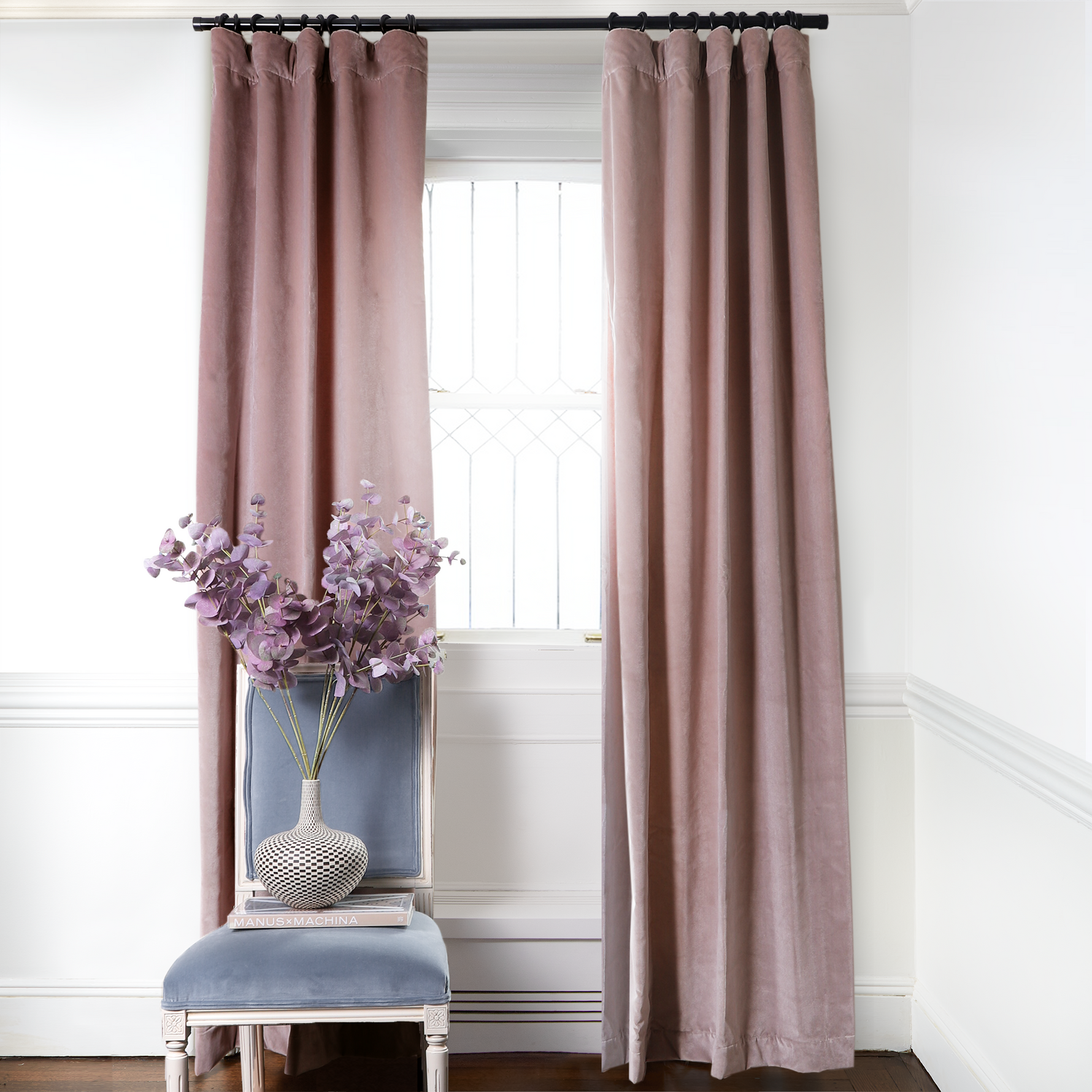 Mauve Velvet Curtains on metal rod in front of an illuminated window with Sky Blue Velvet chair with purple flowers in white and black vase on top book