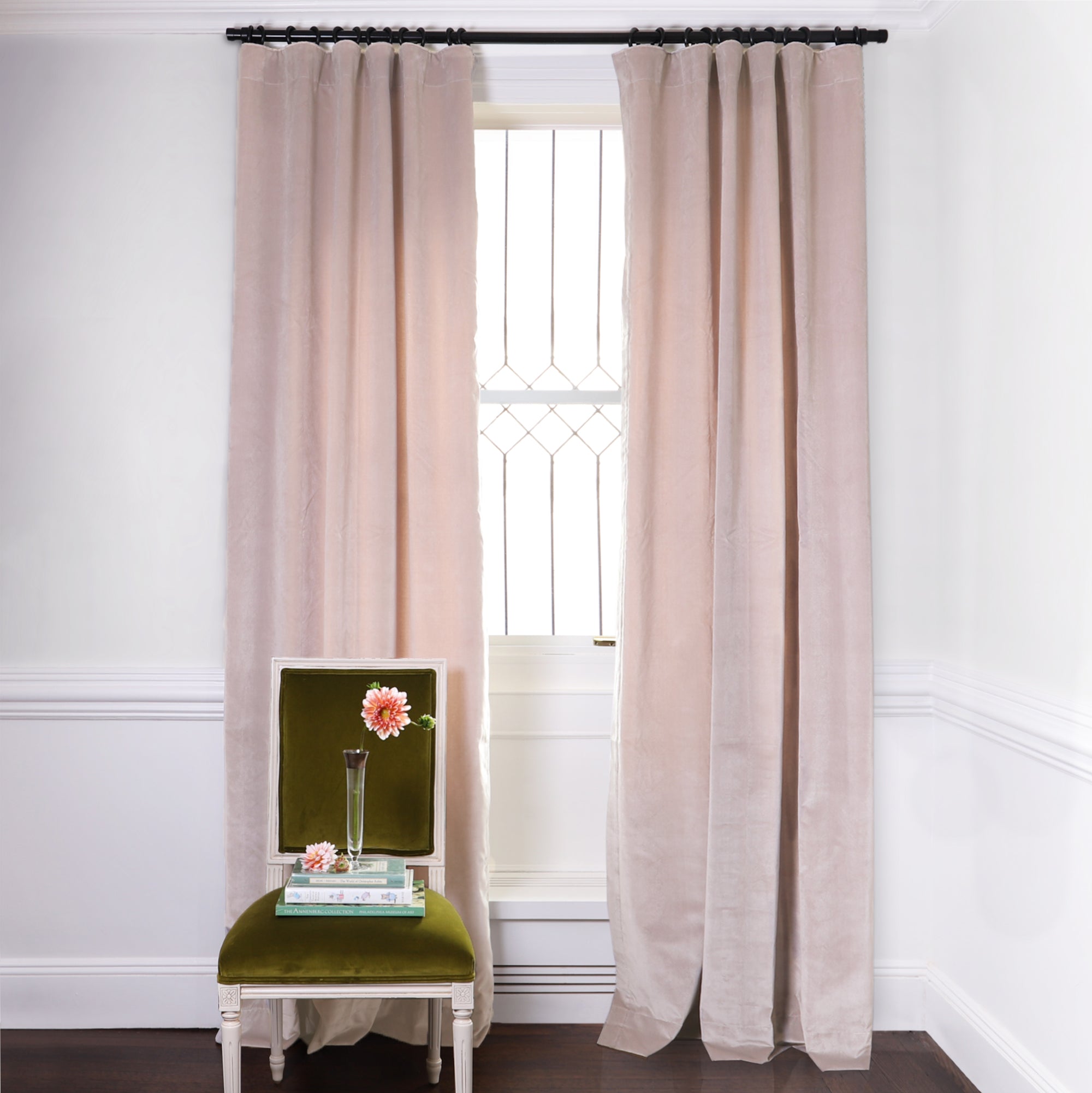 Pink Velvet Curtains on black rod in front of an illuminated window with Moss Green Velvet chair with flowers in clear vase on top of two stacked books