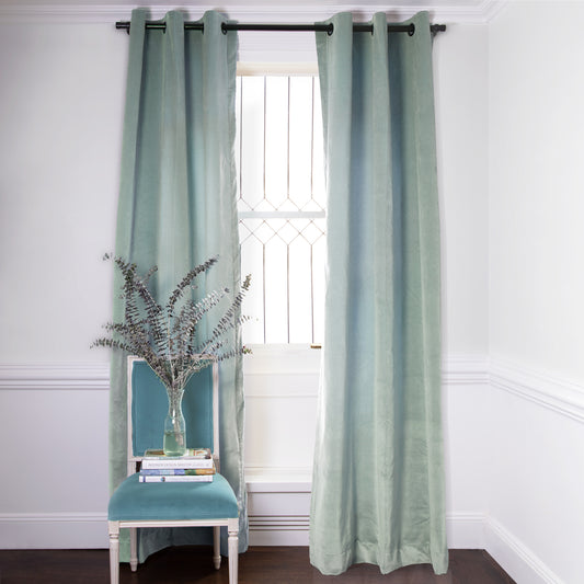 Blue Green Velvet Curtains on black rod in front of an illuminated window with Blue Green Velvet chair with plants in clear vase on top of two stacked books