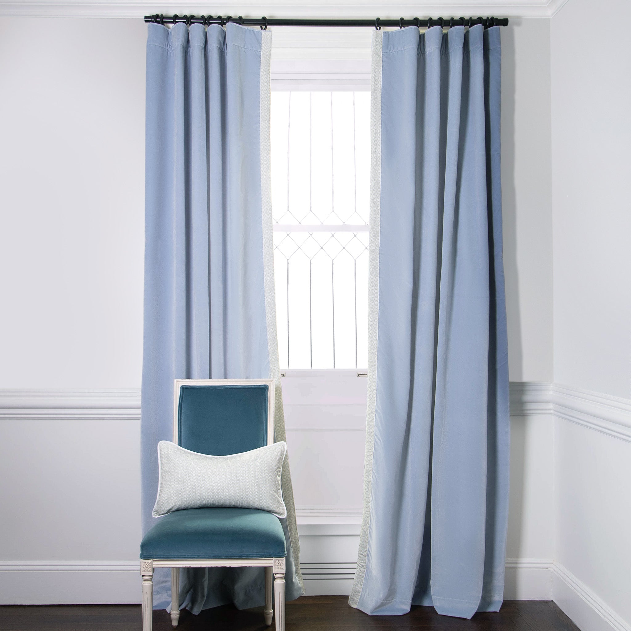 Sky Blue Velvet Curtains on black rod in front of an illuminated window with Blue Velvet chair with a Moss Green Geometric Printed lumbar on top