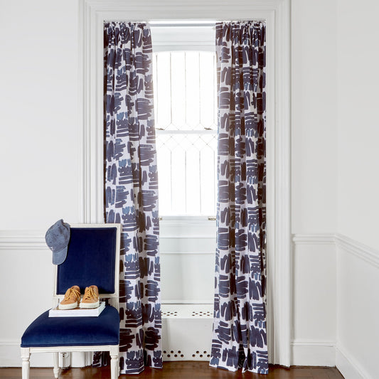 Navy Brushstokes Pattern Custom Curtains on metal rod in front of an illuminated window with a blue navy chair with a kid’s cap hanging from the top and a book and a pair of kid’s shoes stacked on top