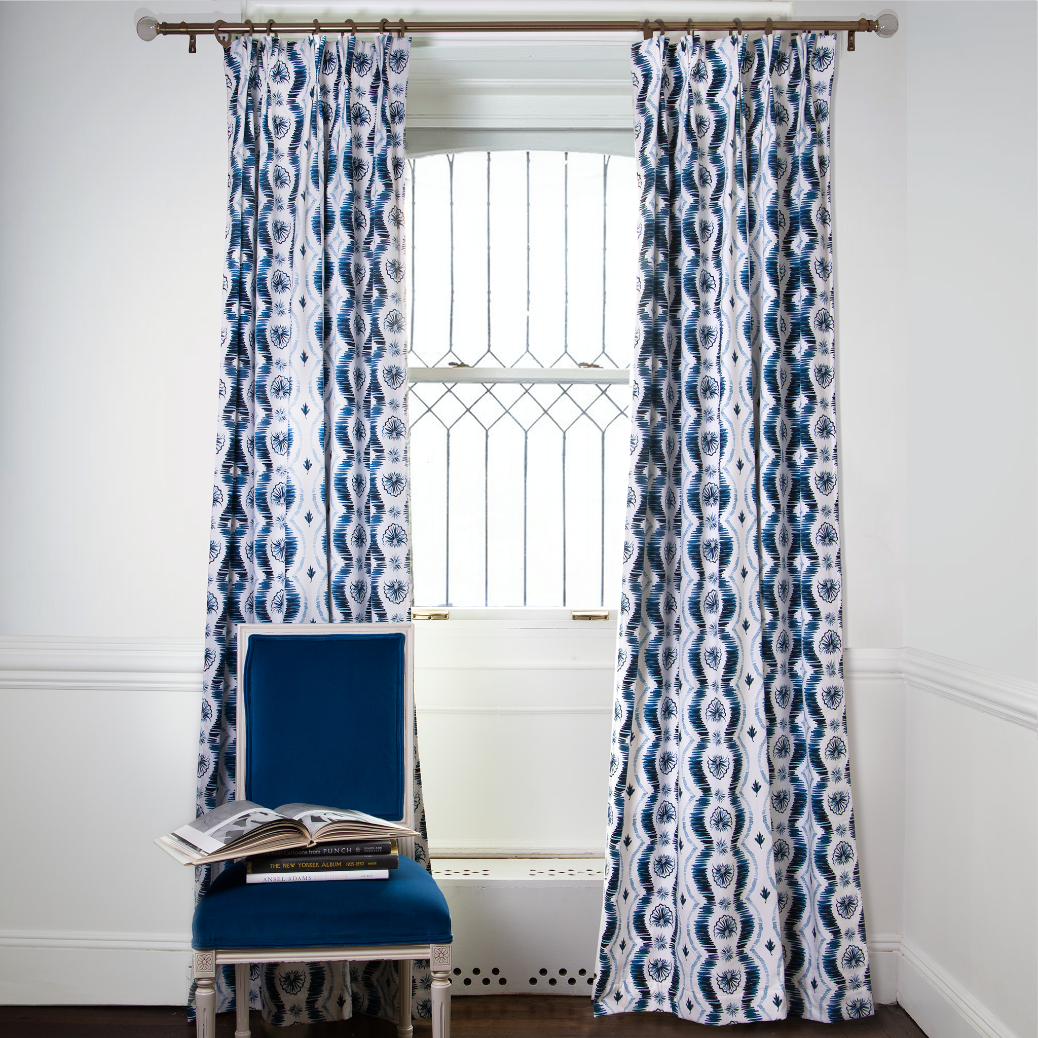 Blue Ikat striped curtains on metal rod in front of an illuminated window with navy chair in front stacked with books