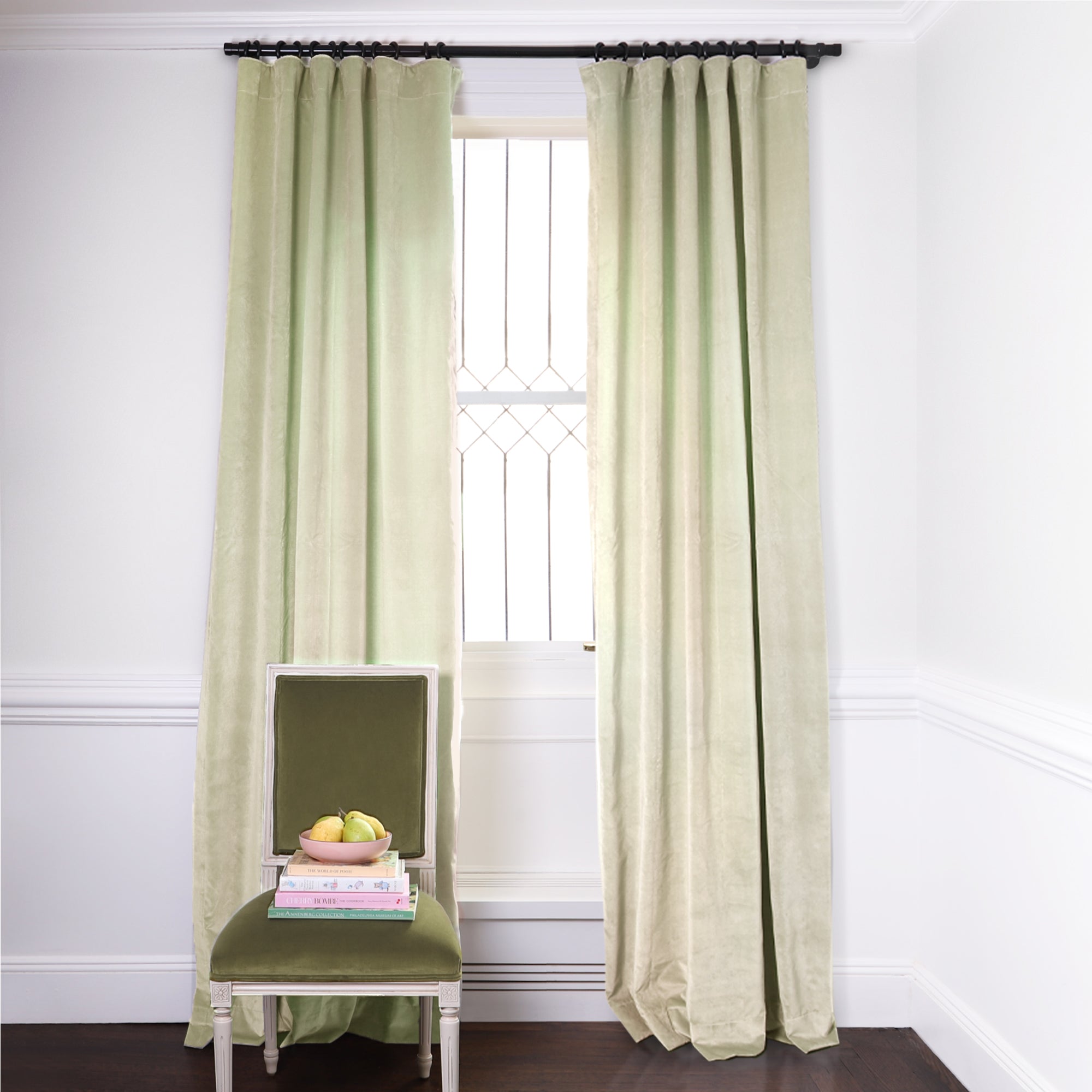 Light Green Velvet Curtains on metal rod in front of an illuminated window with Green Velvet chair with fruit bowl on top of stacked books