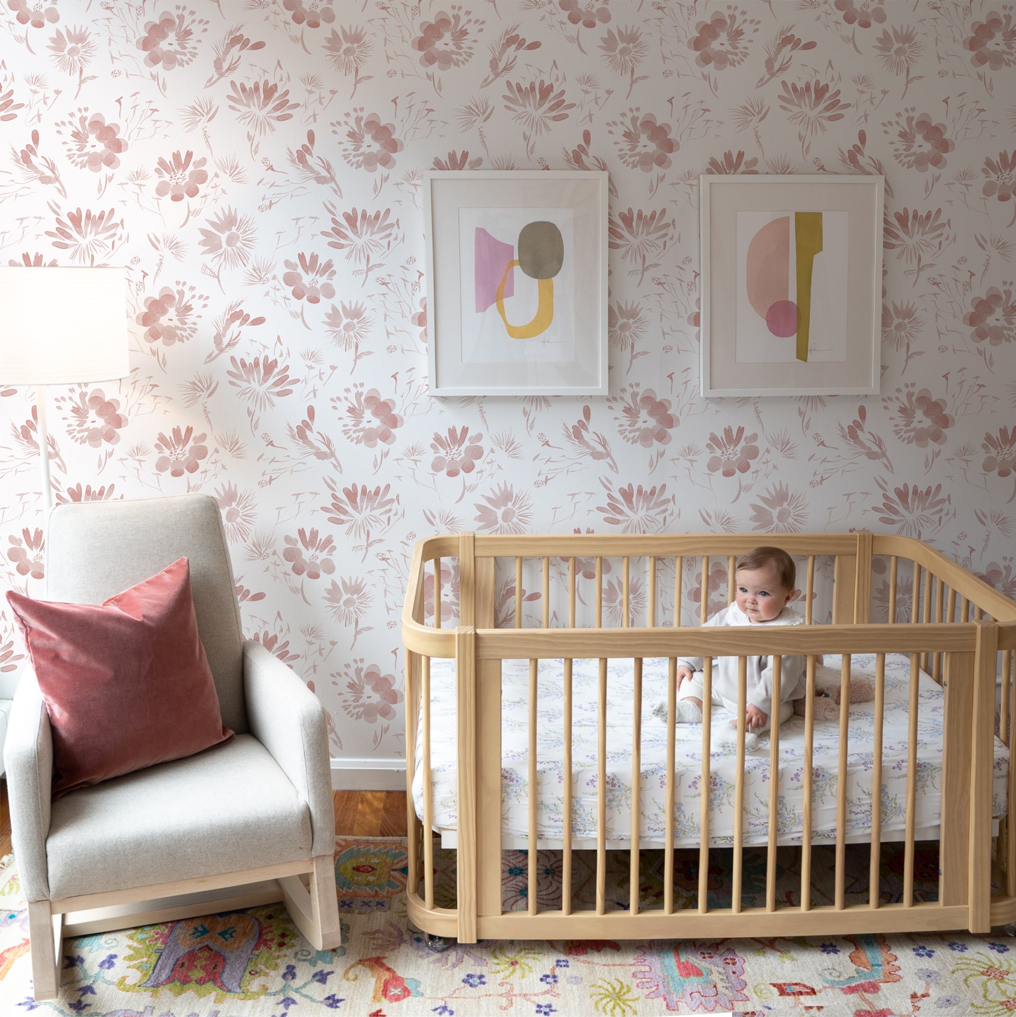 Nursery room with Pink Floral Custom Wallpaper with two art frames on top of crib with baby on top next to White armchair with Coral Velvet Pillow on top next to White tall lamp
