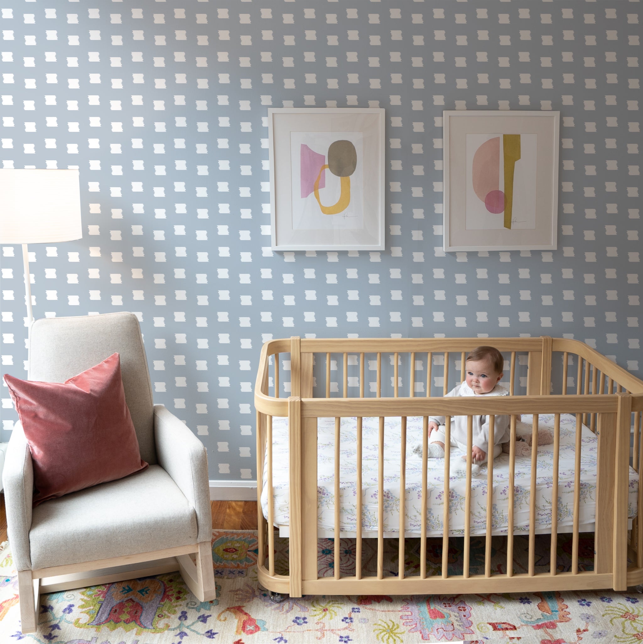 Nursery room with Sky Blue Pattern Wallpaper with two art frames on top of crib with baby on top next to White armchair with Coral Velvet Pillow on top next to White tall lamp