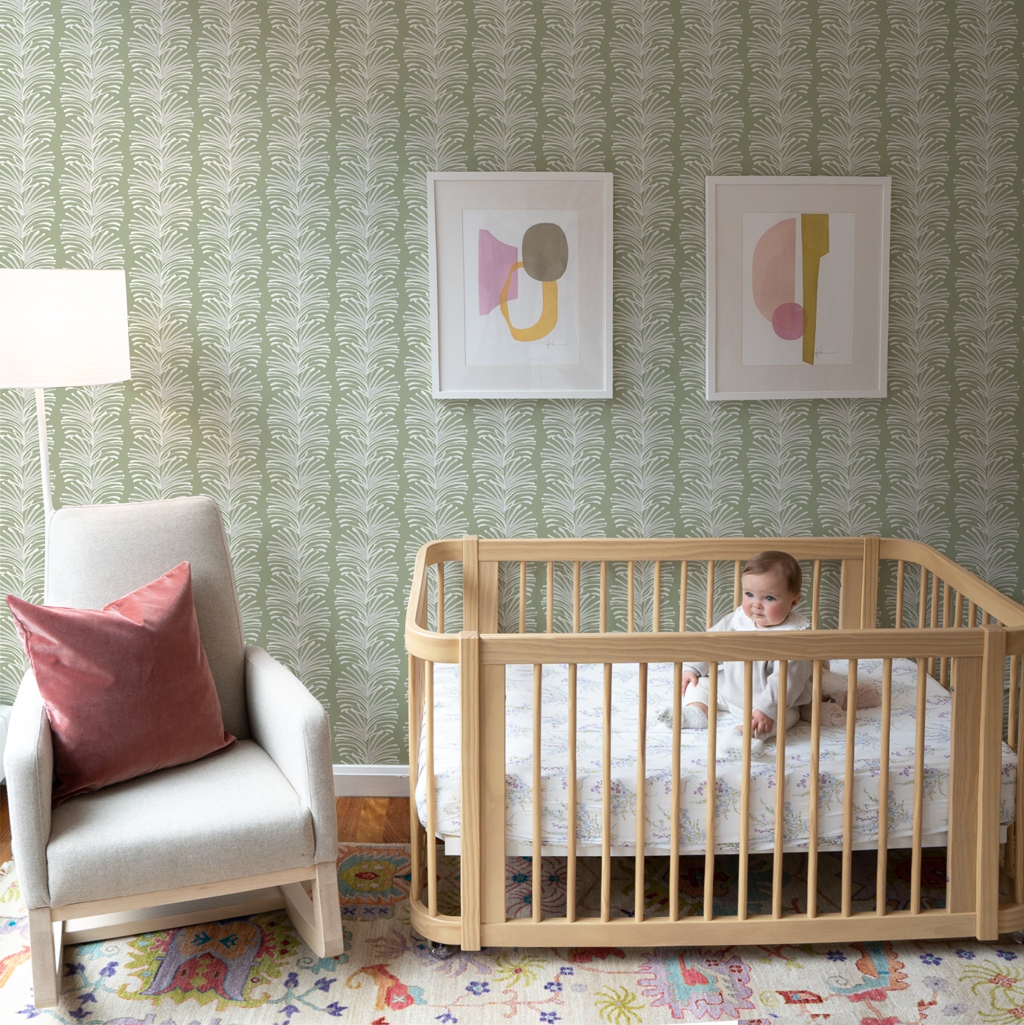 Nursery room with Sage Green Palm Wallpaper with two art frames on top of crib with baby on top next to White armchair with Coral Velvet Pillow on top next to White tall lamp 