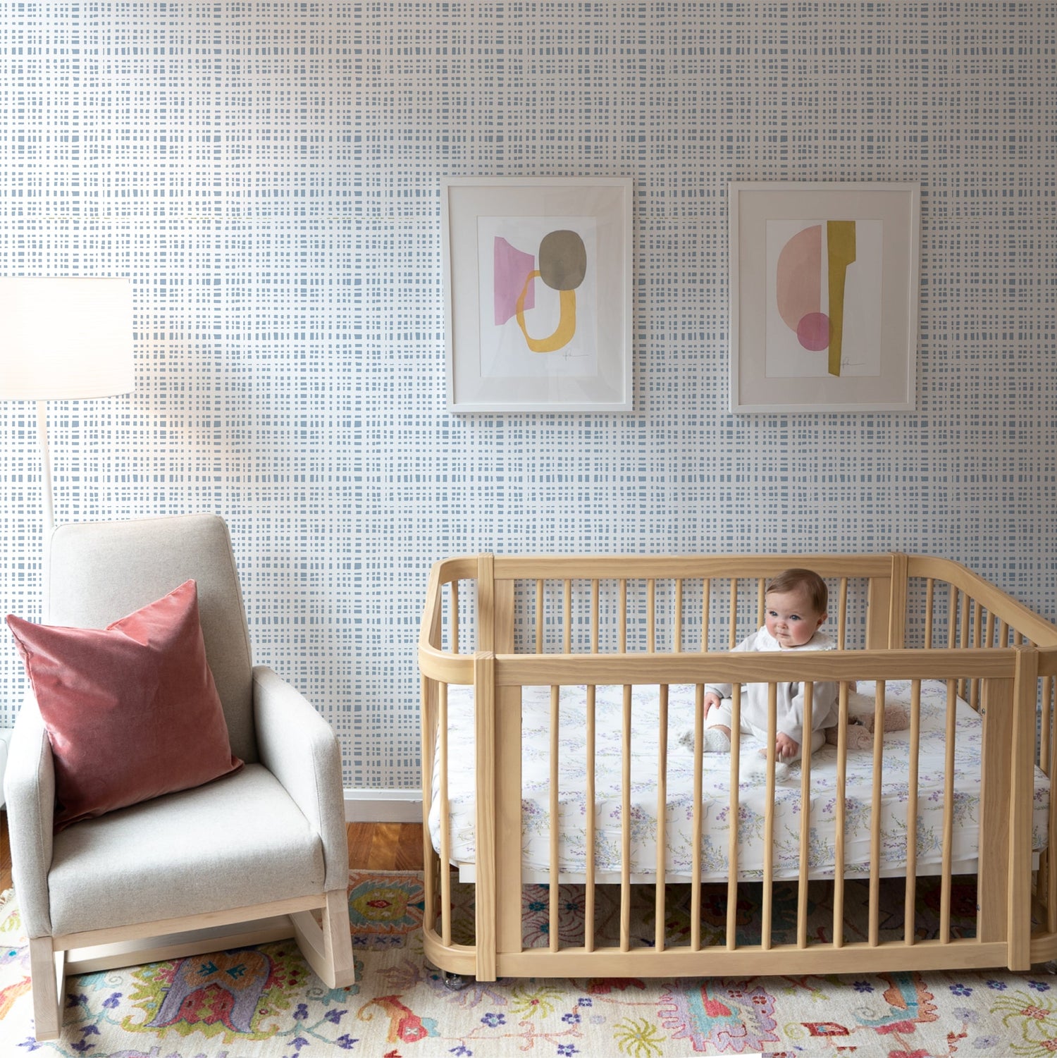 Nursery room styled with Sky Blue Gingham Printed Wallpaper next to wooden crib with baby on it by cream sofa chair with Coral Velvet Pillow on top