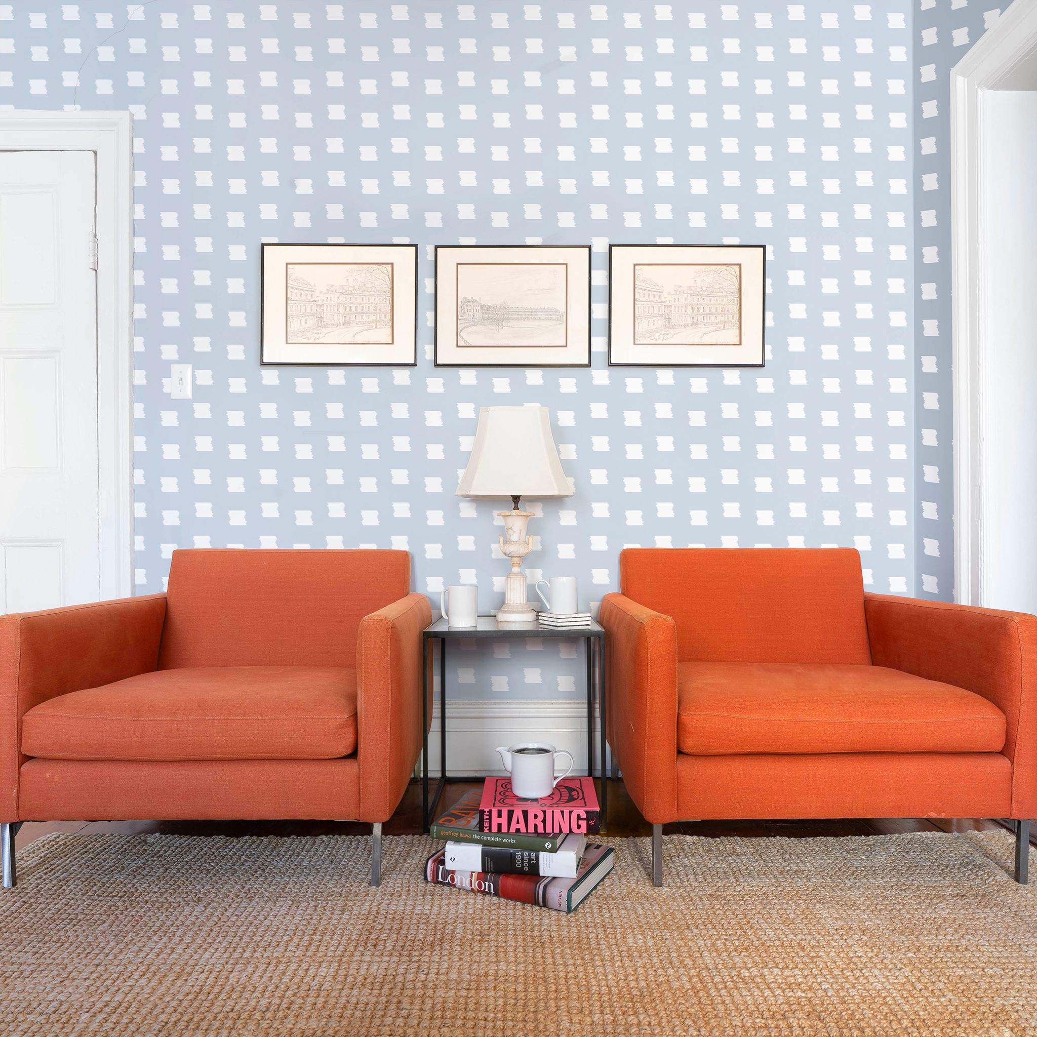 Living room styled with Sky Blue Pattern Printed Wallpaper and three frames over two orange sofa couches with white lamp and decorations on table in between them and stacked books on a light brown rug