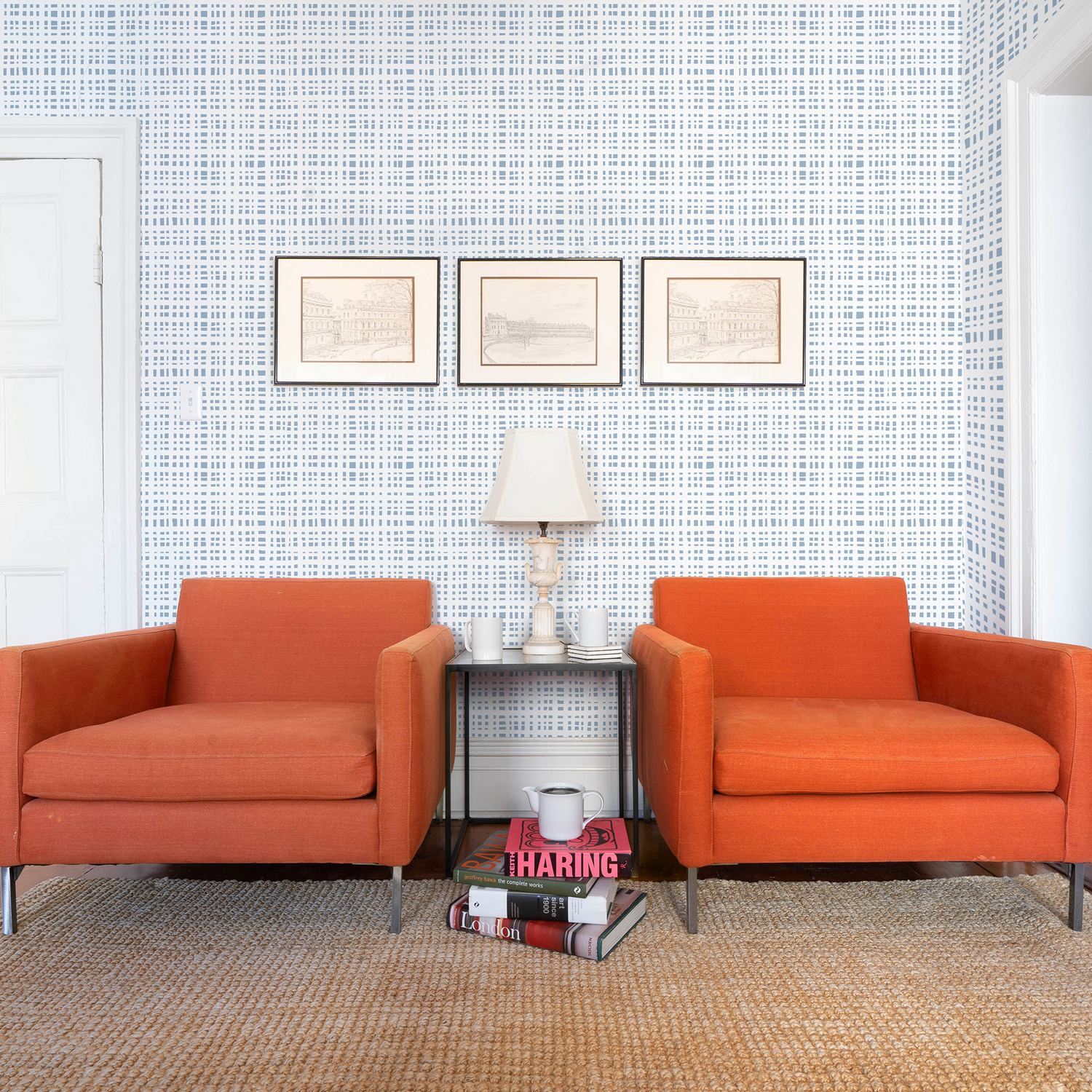 Living room styled with Sky Blue Gingham Printed Wallpaper and three frames over two orange sofa couches with white lamp and decorations on table in between them and stacked books on a light brown rug