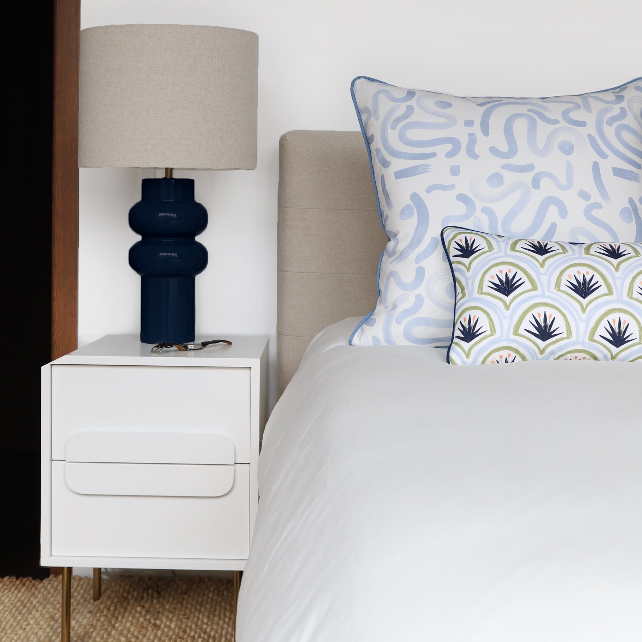 White Bed Close-up styled with a Sky Blue Printed Pillow and an Art Deco Palm Pattern Printed Lumbar next to white nightstand with navy and oat lamp on top