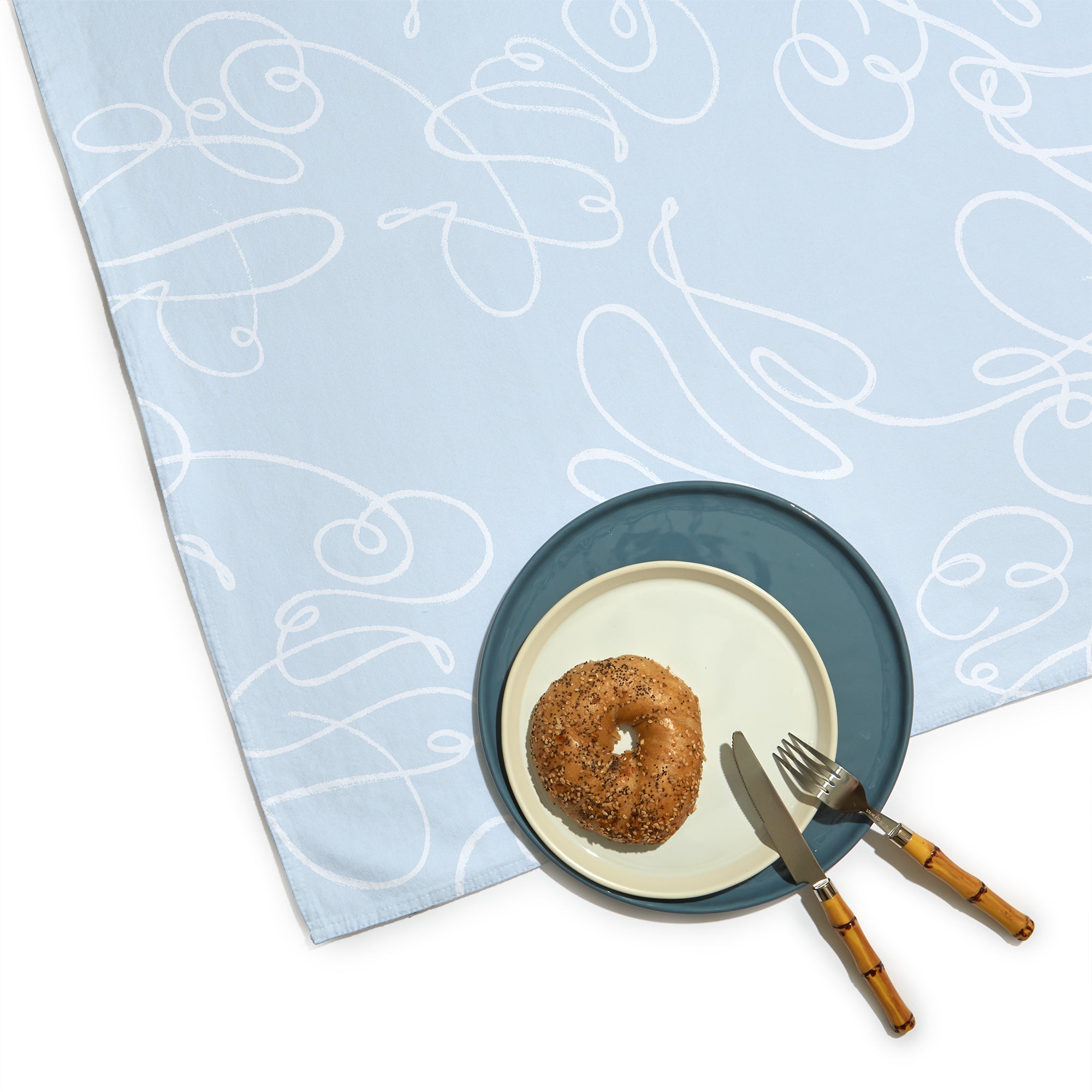 Corner Close-Up of Powder Blue Abstract Printed Table Cloth with bagel on white plate on blue plate with silverware